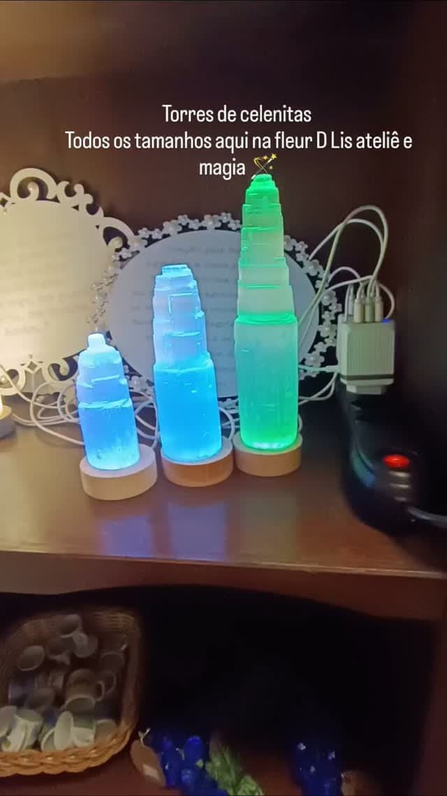 Três torres de selenita iluminadas, uma azul e duas verdes, numa prateleira de madeira